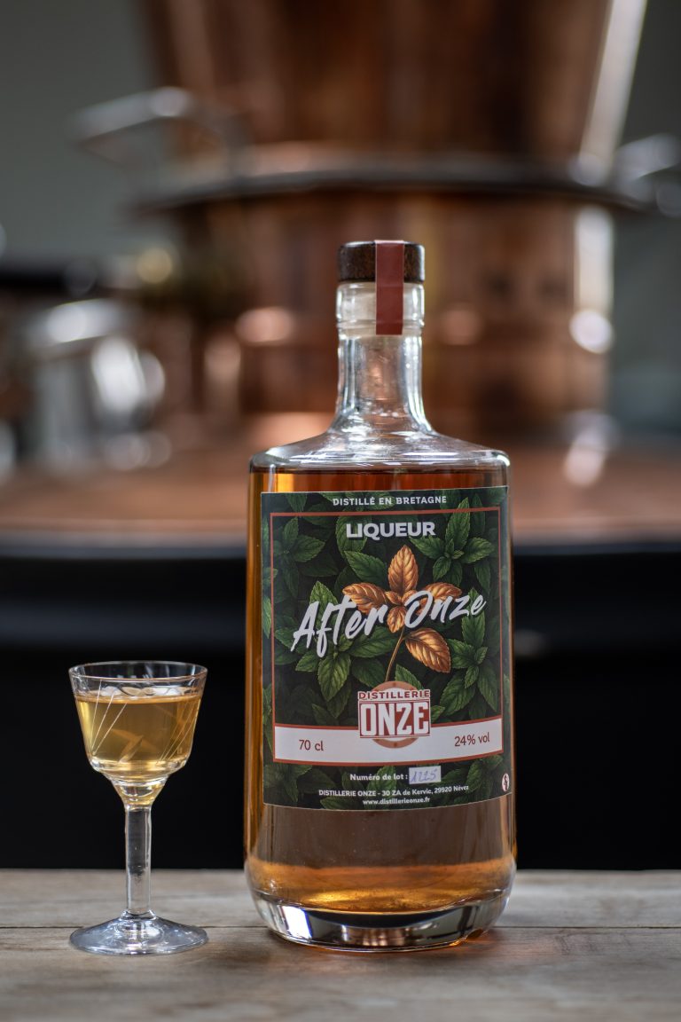 Bouteille de liqueur "After Onze" de la distillerie Onze, posée sur une table en bois à côté d'un verre à dégustation rempli.